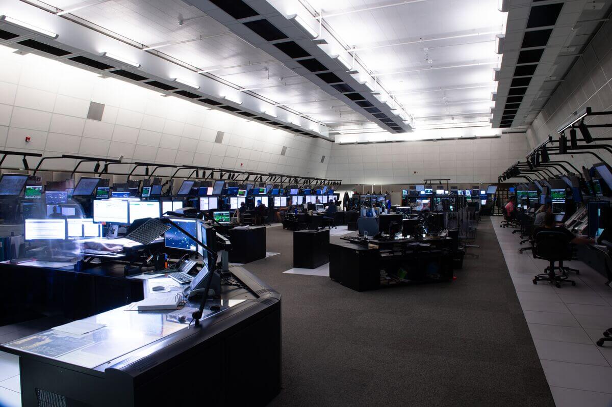 Interior view of a modern area control centre. The room is dimly lit, primarily by the glow of computer screens. Multiple rows of desks are equipped with computer monitors. 